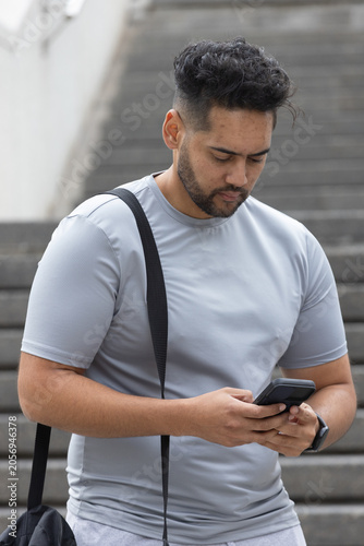 African American man standing on concrete steps, checking phone, carrying bag strap, wearing watch