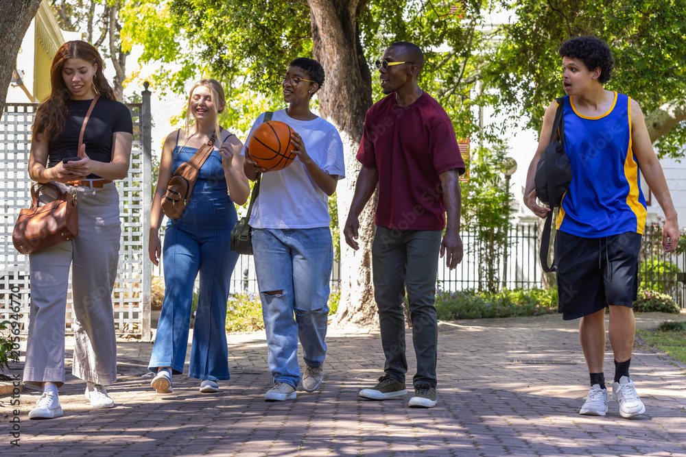 Naklejka premium Diverse friends walking along shaded brick path, carrying basketball, bags, wearing sporty clothes