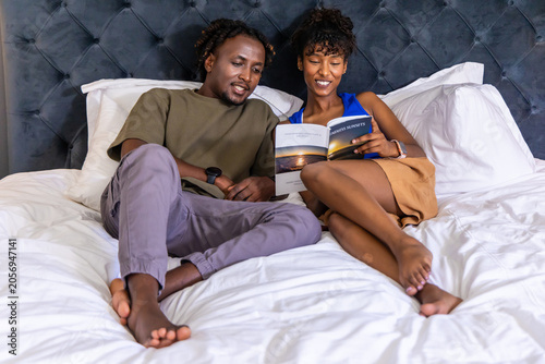 African American couple reclining on bed reading book with sunset picture, wearing wristbands