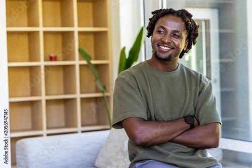 Quadro su tela Man sitting on light-colored sofa, crossing arms, smiling near wooden cubby with