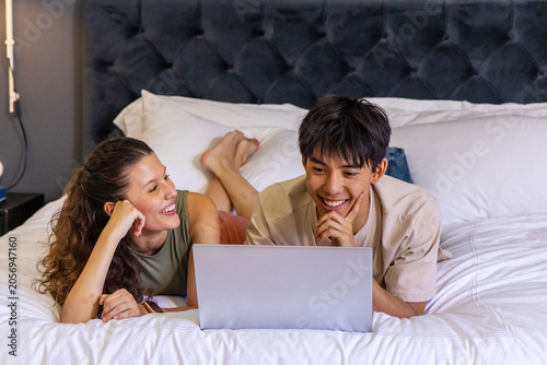 Couple lying on stomachs on large bed using silver laptop in contemporary bedroom