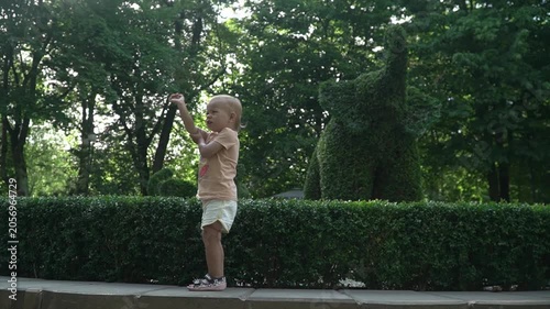 A small child, girl, points figure elephant from bush, park with landscaping. On your trunk and on your nose. Laughs, smiles, jumps. In shorts, T-shirt, sandals, with short hair. Day, summer