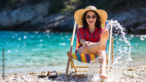 Joyful woman in straw hat and red plaid shirt splashing water with feet on pebble beach, summer vacation lifestyle.