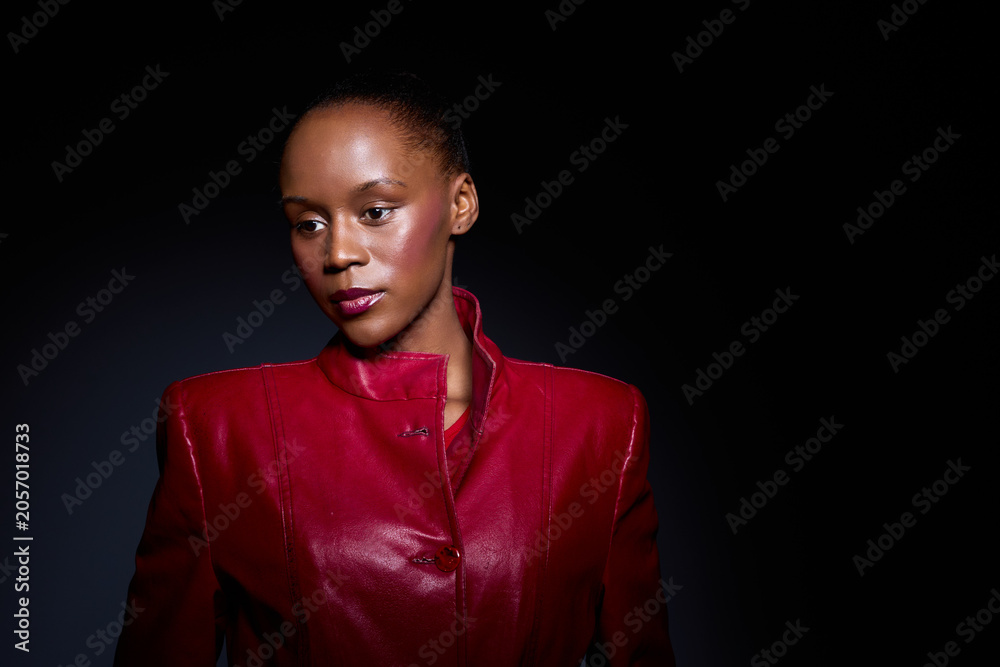 Obraz premium Elegant dark skinned African woman in red jacket poses against black background. Studio portrait with dramatic lighting highlights her confident expression and fashion style.