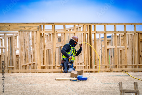 Construction worker holding air hose and pneumatic nail gun while standing on OSB roof of wood-framed building