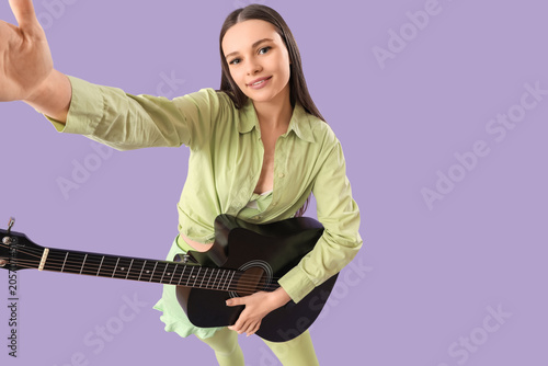 Young girl with guitar taking selfie on lilac background