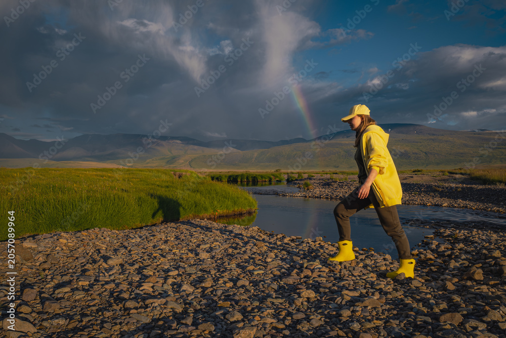 Fototapeta premium Side view of young woman in yellow rain boots hiking near creek with rainbow in sky. Female traveler exploring remote mountain valley landscape after storm during golden hour.