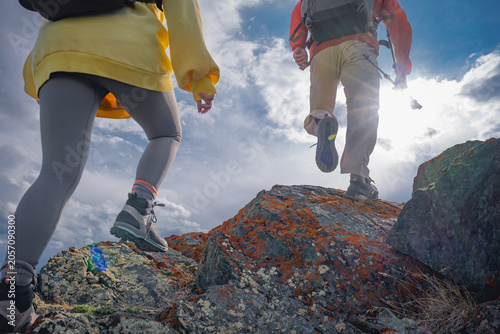 Low angle rear view of young couple hiking up rocky mountain hill. Active man and woman climbing peak under beautiful cloudy sky with bright sun flare during expedition.