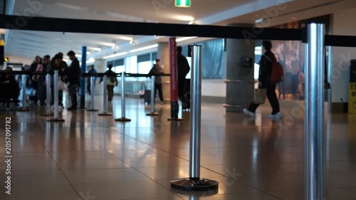 Close-up on a chrome retractable belt queue barrier stanchion in foreground of a busy airport terminal, with blurred passengers and travellers moving through concourse in a busy international airport