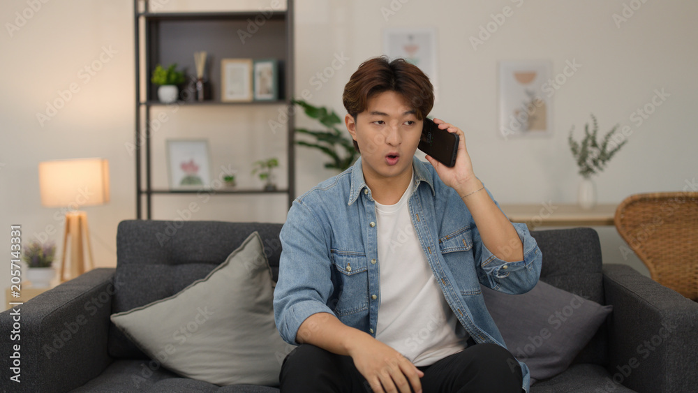 Obraz premium Young Asian Man with Surprised Expression Talking on Smartphone in Living Room