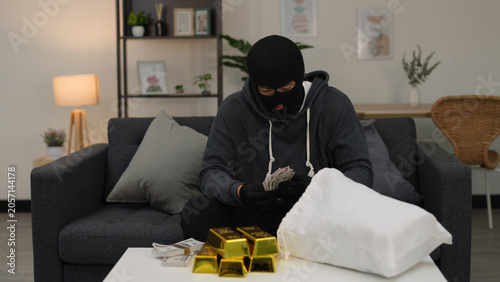Photos Man in Balaclava Counting Cash and Gold Bars in Living Room
