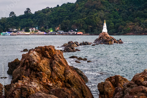 Scenic View of White Ban Hua Laem Pagoda on Rocky Island. The white Ban Hua Laem Pagoda situated on a rocky island in the sea, with lush coastline, local pier, and calm water in Chanthaburi, Thailand.