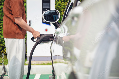 Person plugs charging cable into an electric car. They wear brown sweater and light trousers. Charging station has white structure with green trees in background. Signifying the use of clean energy.