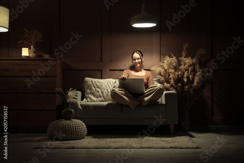 Smiling young woman using laptop on sofa at night, remote work and online lea...