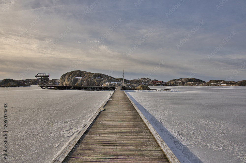 Obraz premium Symmetrical view of a wooden pier with rope railings leading across the ice in Skrhamn. A cold winter day in the Swedish archipelago with dramatic light and frozen wate