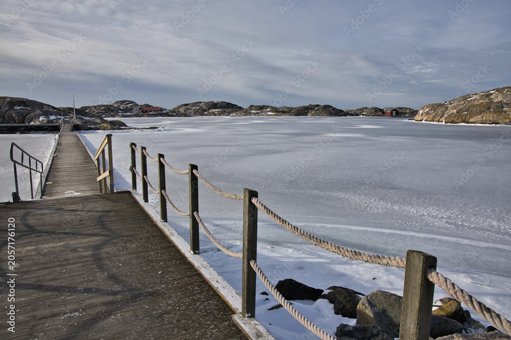 Naklejka premium Symmetrical view of a wooden pier with rope railings leading across the ice in Skrhamn. A cold winter day in the Swedish archipelago with dramatic light and frozen wate