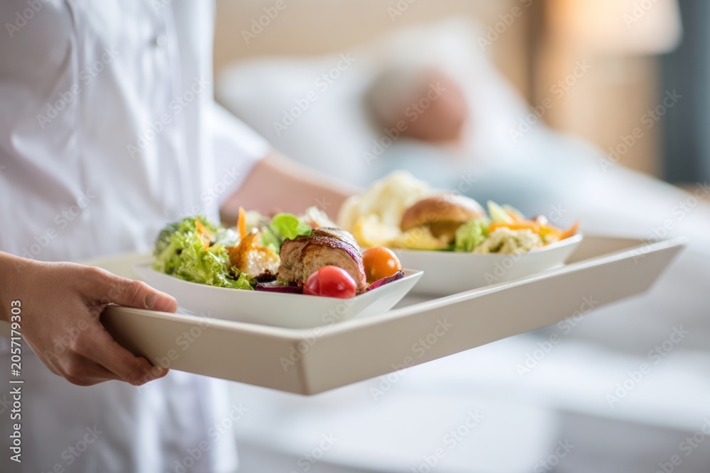 Naklejka premium A young adult female caregiver with fair skin in a white uniform carries a nutritious food tray to a patient resting in a bright hospital room.