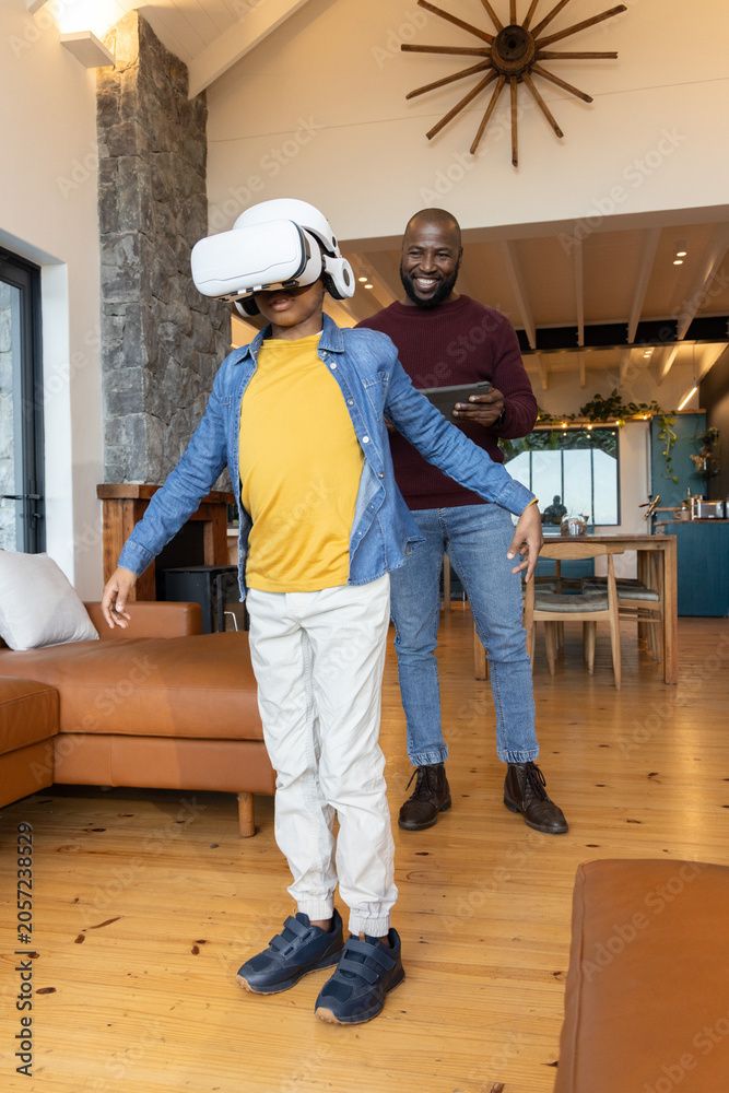 Naklejka premium African American father and son playing in living room child wearing VR headset father using tablet