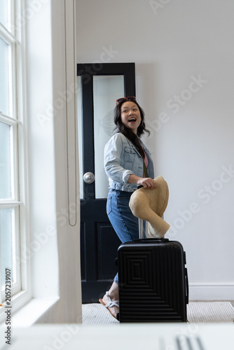Asian woman standing in bright entryway holding straw sun hat and gripping black suitcase handle