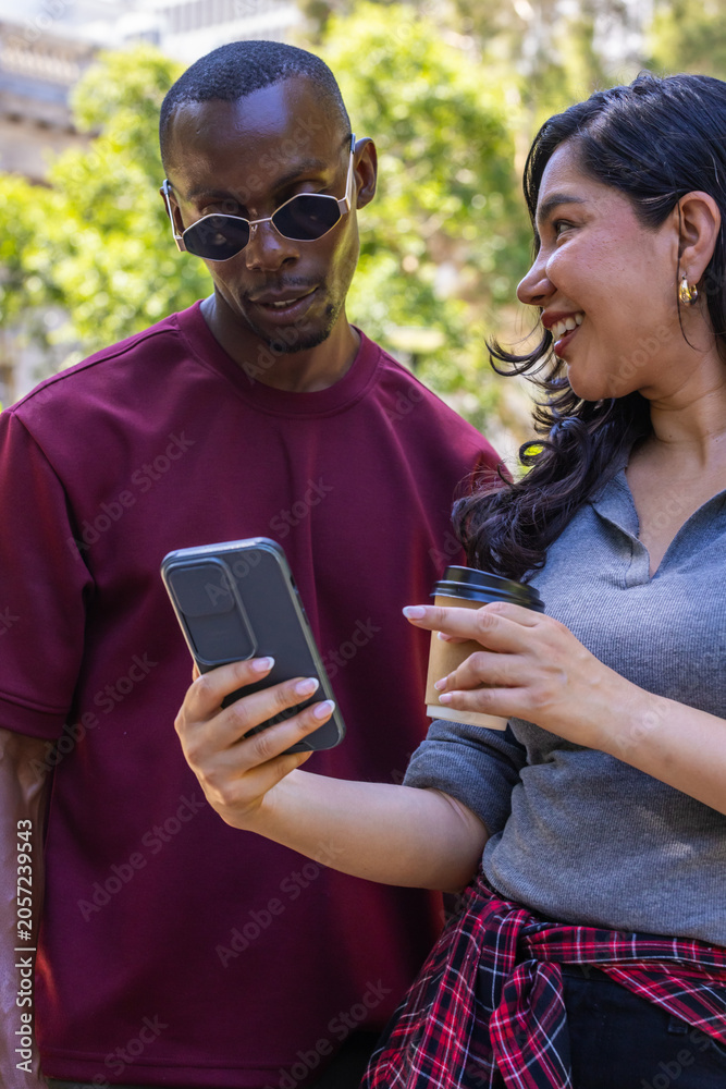 Fototapeta premium Diverse couple leaning showing phone holding coffee wearing shades and plaid tied at waist in plaza