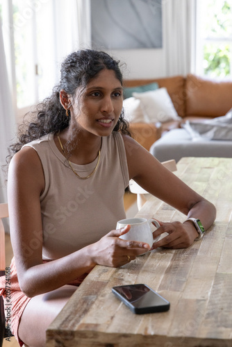 Adult Indian woman in sleeveless beige top sitting at table at home holding white mug, smartphone