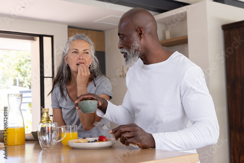 Diverse couple sitting at kitchen island tasting pancakes with berries near carafe of orange juice