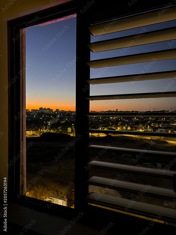 Fototapeta premium Cityscape view through window blinds at twilight, urban horizon with glowing lights and sunset sky, the sunset sky displays a gradient of orange and blue hues