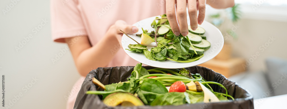 Naklejka premium Person scraping leftover vegetable salad from plate into a trash bin, food waste concept.