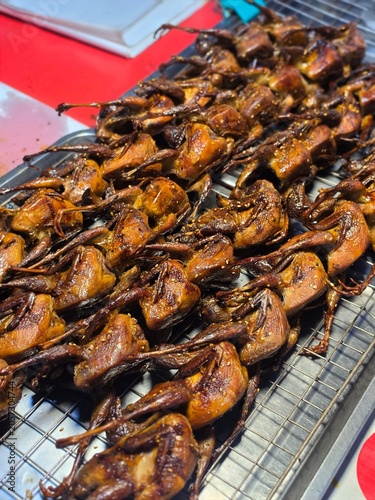 Roasted poultry quail in marinade seasoning or grilled partridge marinated in spices on machine gas stove for thai people eat in street market bazaar annual event festival in Nonthaburi city Thailand