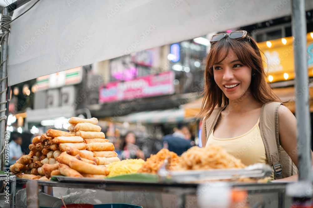 Fototapeta premium Young woman enjoy eating street food at night market. Traveler blogger women Happy tourists Beautiful female with Traditional food local road at thailand bangkok city.