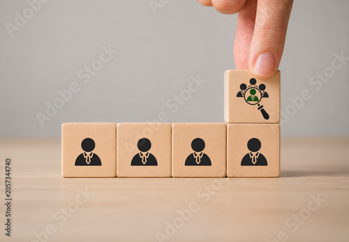 Recruitment, team building and talent acquisition in a business. Wooden blocks displaying icons of employees, with a hand placing a block showing a magnifying glass highlighting a selected individual
