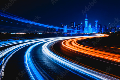 Long exposure shot showing light trails from moving vehicles on a highway at night, with a lit city skyline in the background.