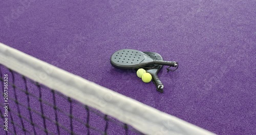 Camera starting and showing padel rackets and tennis balls resting on purple court behind net
