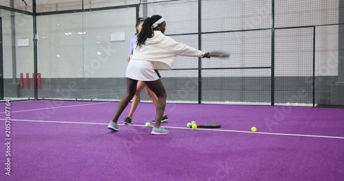 Black student practicing forehands with racket as indian coach feeding balls on padel court