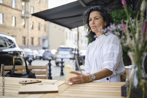 A woman sits at a wooden table outside a cafe, holding a glass of cold lemonade. She has dark, wavy hair and is wearing a white blouse