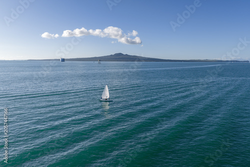 A lone sailboat navigates the waters of the Hauraki Gulf near Auckland, New Zealand, with Rangitoto Island in the background.