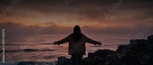 Silhouetted figure with arms outstretched standing on rocky coastline at sunset, experiencing freedom and contemplation by the sea during golden hour.
