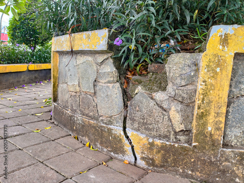 Damaged stone planter wall with deep crack and peeling yellow paint. Broken concrete retaining barrier for green plants on paving block sidewalk background.