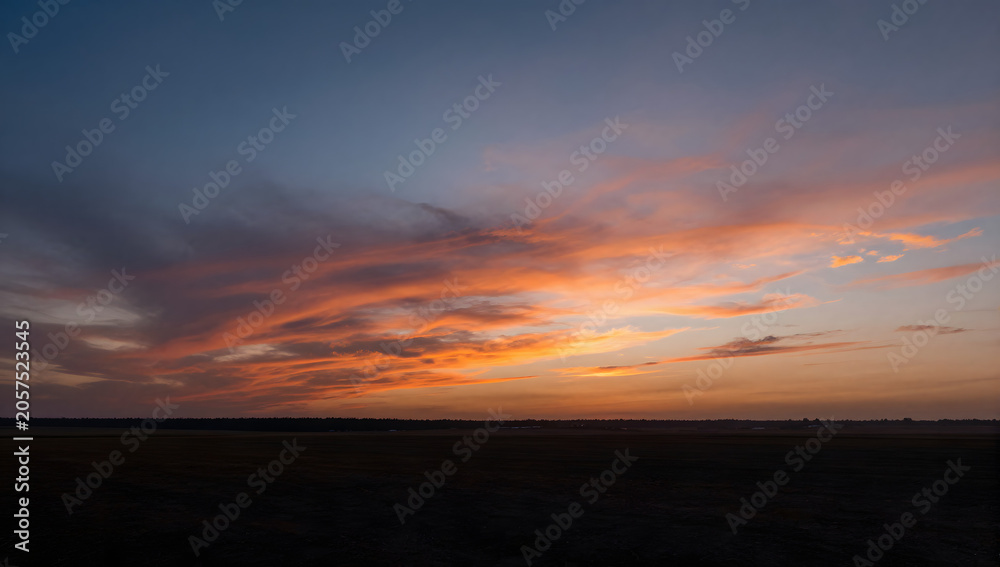Fototapeta premium Dramatic Sunset Over Open Field: The sky explodes with vibrant hues of orange and pink as a fiery sunset paints the clouds above a vast, dark expanse of a field.
