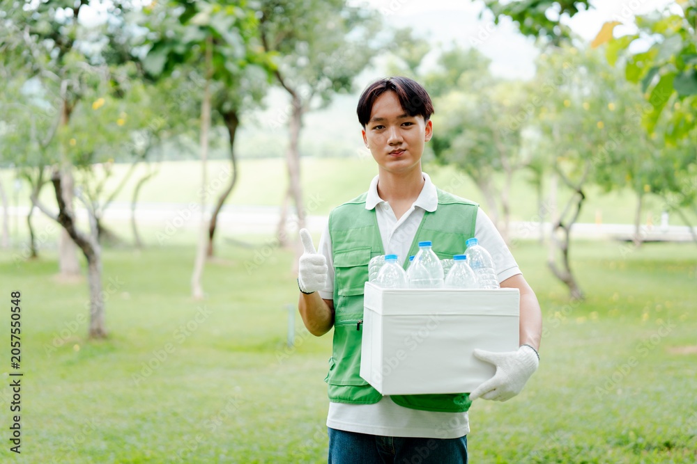 Fototapeta premium Young asian man volunteer wearing green vest and gloves. Holding a box of plastic bottles for recycling. Proudly giving thumbs up gesture in a park