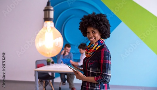 Creative portrait of a young woman with afro hair holding a tablet in an office with a large glowing lightbulb, innovation