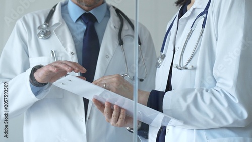 Female doctor writing on clipboard while male colleague discusses patient data beside her, illustrating teamwork, consultation and coordinated care in a clinical setting