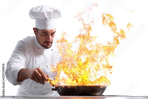 A focused chef in a white uniform and toque expertly cooks with flames erupting from a pan, showcasing a dramatic culinary moment.