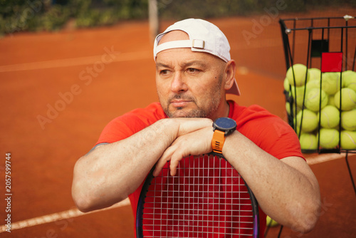 Tennis coach holding a racket on a clay court, professional sports setting with focused confident posture, natural daylight