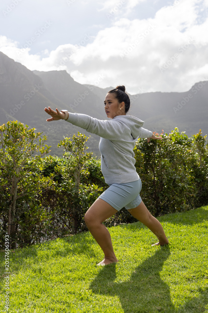 Fototapeta premium Standing lunge pose balancing on well kept grassy lawn, with trimmed hedge and mountain ridge