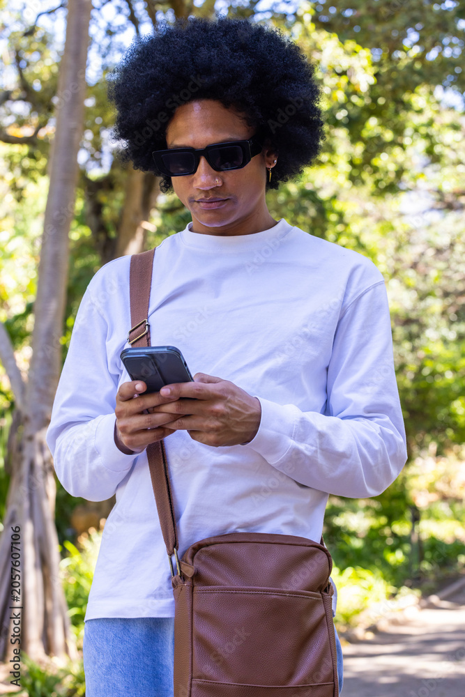 Fototapeta premium African American adult man standing, checking smartphone, wearing shirt and crossbody bag in park