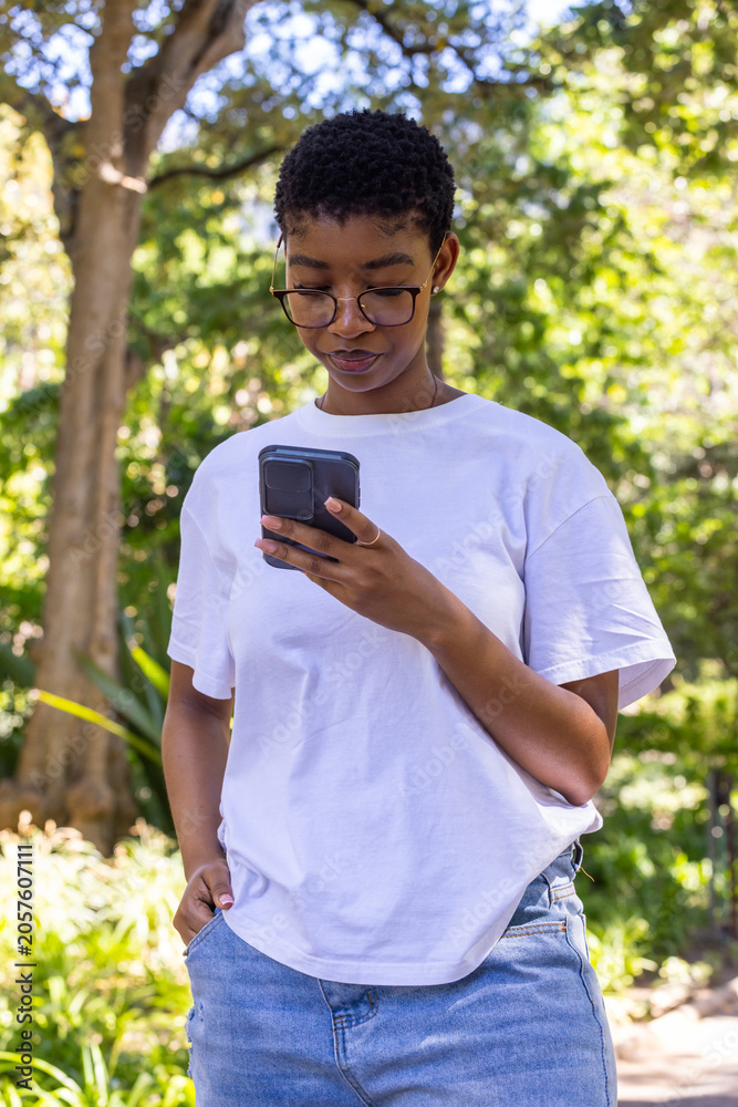 Fototapeta premium African woman standing in sunny park wearing white tee and jeans wearing glasses holding smartphone