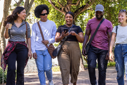 Diverse friends walking and chatting on brick pathway in park, showing smartphone and coffee