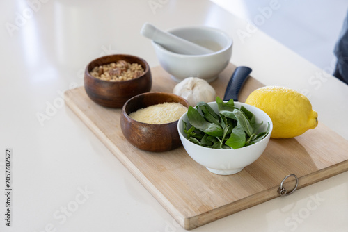 Wooden cutting board is holding basil bowl, cheese, pine nuts, garlic, lemon on counter, copy space