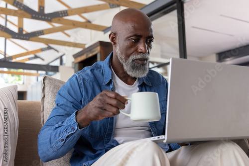 Mature African American man sitting on sofa, wearing denim shirt, holding mug while using laptop